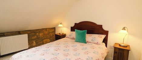 A bedroom with a sloped ceiling, a stone wall, a wooden headboard, a floral bedspread, and two bedside lamps.