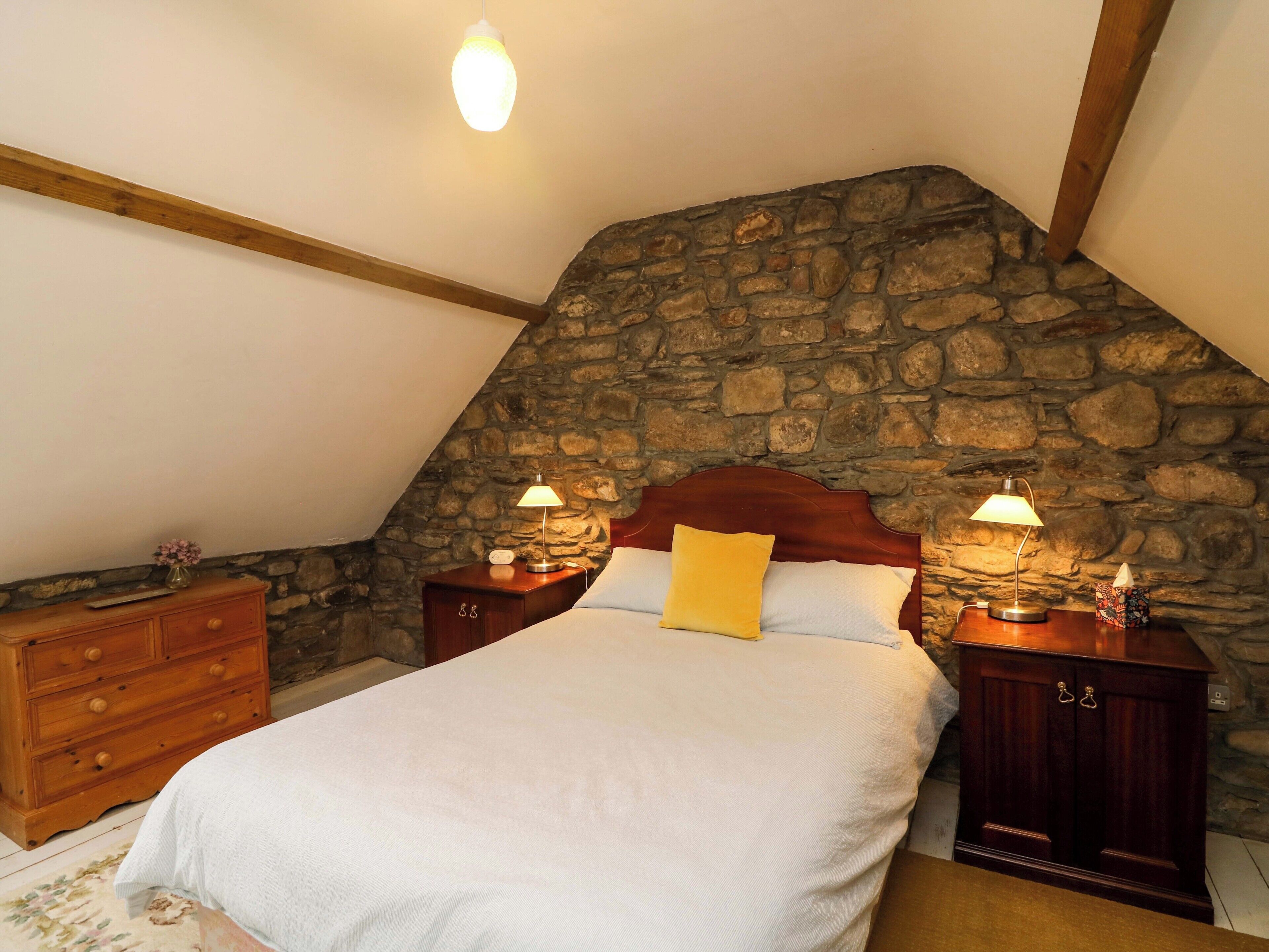 A bedroom with a stone wall, wooden headboard, a bed with white bedding, two bedside tables with lamps, and a wooden dresser.