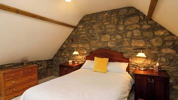 A bedroom with a stone wall, wooden headboard, a bed with white bedding, two bedside tables with lamps, and a wooden dresser.