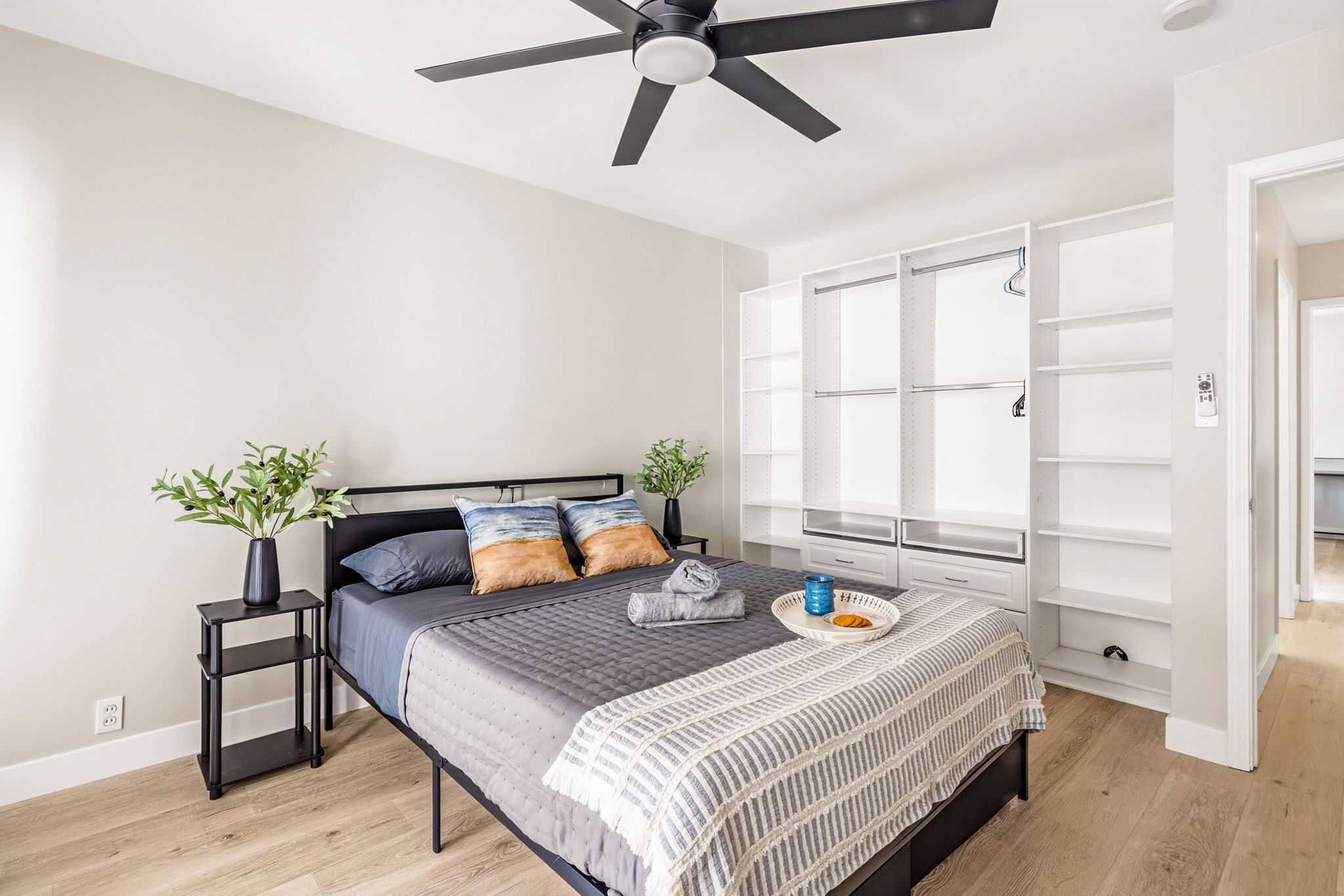 A bedroom with a bed, a ceiling fan, a nightstand, a potted plant, a window with blinds, and a built-in wardrobe.