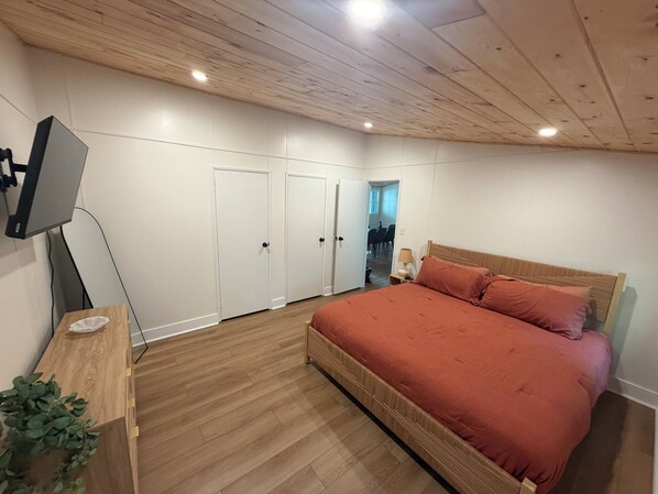 Um quarto com teto de madeira, uma cama com colcha vermelha, uma mesinha de cabeceira e uma televisão fixada na parede.
