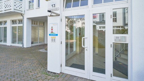 Ein weißes Gebäude mit der Hausnummer 13, einem Glastür-Eingang und einem Balkon mit weißer Brüstung.