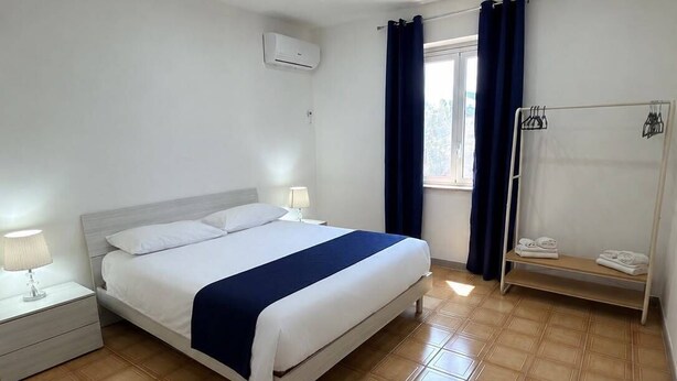 Quarto de hotel com cama, mesa de cabeceira, janela com cortinas, porta-toalhas e ar-condicionado fixado na parede.