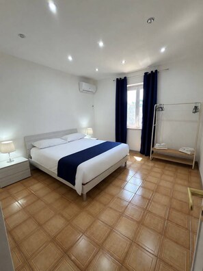 Une chambre d’hôtel comprenant un lit, une table de chevet, une fenêtre avec des rideaux, un porte-serviettes et un climatiseur fixé au mur.