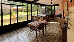 A spacious kitchen with large windows, a dining area with a patterned tablecloth, and a tiled floor.