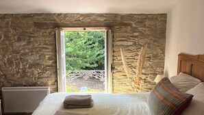 A bedroom with a stone wall, a bed with pillows, a radiator, and a view through a glass door.