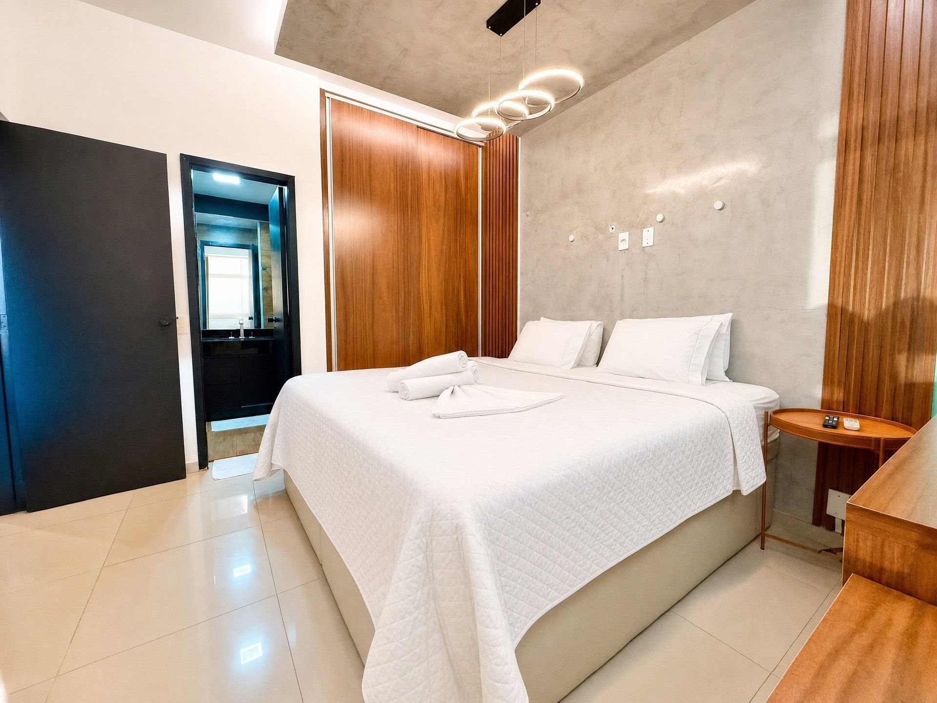 A modern bedroom with a large bed, wooden paneling, a bedside table, and a bathroom visible through an open door.