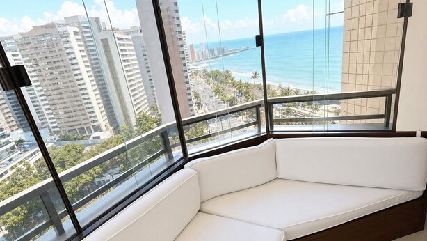 A modern balcony with a white cushioned seating area, large windows offering a view of the cityscape and ocean. - Flat Beira mar com Linda Vista e Hidromassagem (Fortaleza)