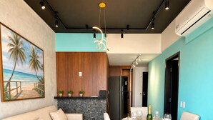 A modern living room with a sofa, dining table, and chairs, a kitchen area with a refrigerator, and a painting on the wall. - Flat Beira mar com Linda Vista e Hidromassagem (Fortaleza)