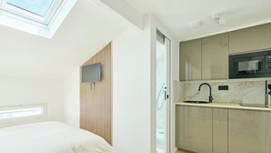 A compact bedroom with a skylight, a bed with a striped blanket, and a kitchenette with a microwave and sink. - Richard Lenoir · 11th · Premium Rooftop Studio – managed by Mey Agency (Paris)