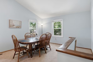 Une salle à manger avec une table et des chaises en bois, deux fenêtres donnant sur des arbres et un tableau accroché au mur.