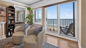 Een woonkamer met een beige bank, een boekenkast en een grote schuifglazen deur die leidt naar een balkon met uitzicht op zee.