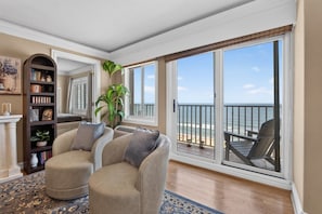 Ein Wohnzimmer mit einem beigen Sofa, einer Bücherwand und einem großen Schiebeglastür, die auf einen Balkon mit Meerblick führt.