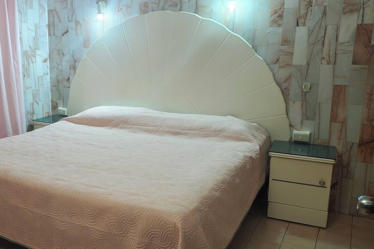 Una cama con cabecera, una mesita de noche, un cajón y una cortina.
