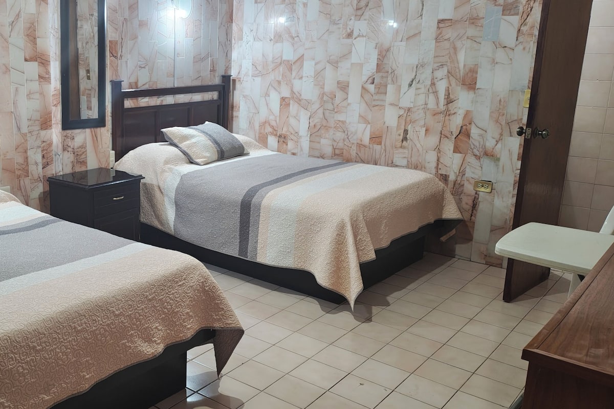 Habitación de hotel con dos camas, una mesita de noche, un banco y un espejo.