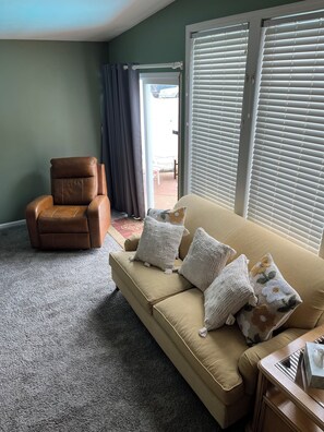 Un salon avec un canapé beige, un fauteuil en cuir marron et une grande fenêtre équipée de stores.