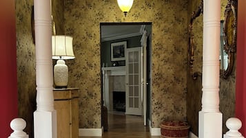Un couloir doté d’un plancher en bois, de colonnes blanches, d’un lustre, d’une lampe et d’une porte donnant sur une autre pièce.