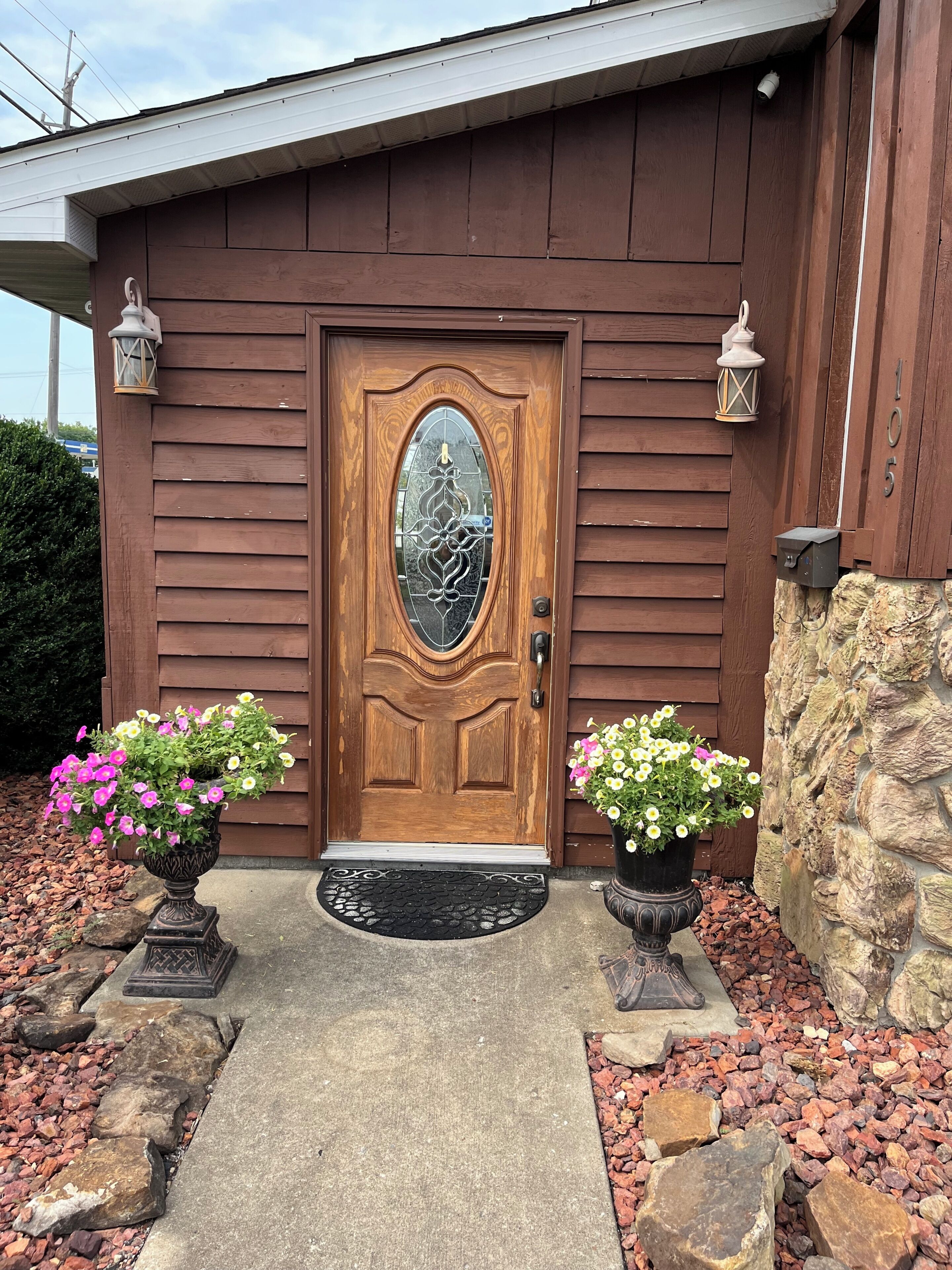 A wooden house with a brown door, two potted plants, a stone wall, and a mailbox.