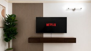 Moderni olohuone, jossa on seinälle asennettu televisio, jossa näkyy Netflix, puinen mediakeskus, ruukkukasvi ja pyöreä puupöytä.