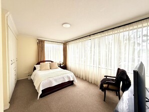 Ein Hotelzimmer mit einem Bett, einem Sessel, einem Schreibtisch, einem Fernseher und großen Fenstern mit Vorhängen.