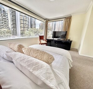 Ein Hotelzimmer mit einem großen Bett, einem Fernseher, einem Sessel und Blick auf hohe Gebäude.