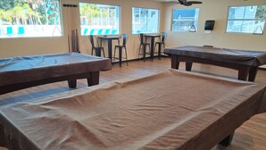 Una sala con mesas de billar, dos mesas y sillas, un ventilador de techo y ventanas con vistas a palmeras.