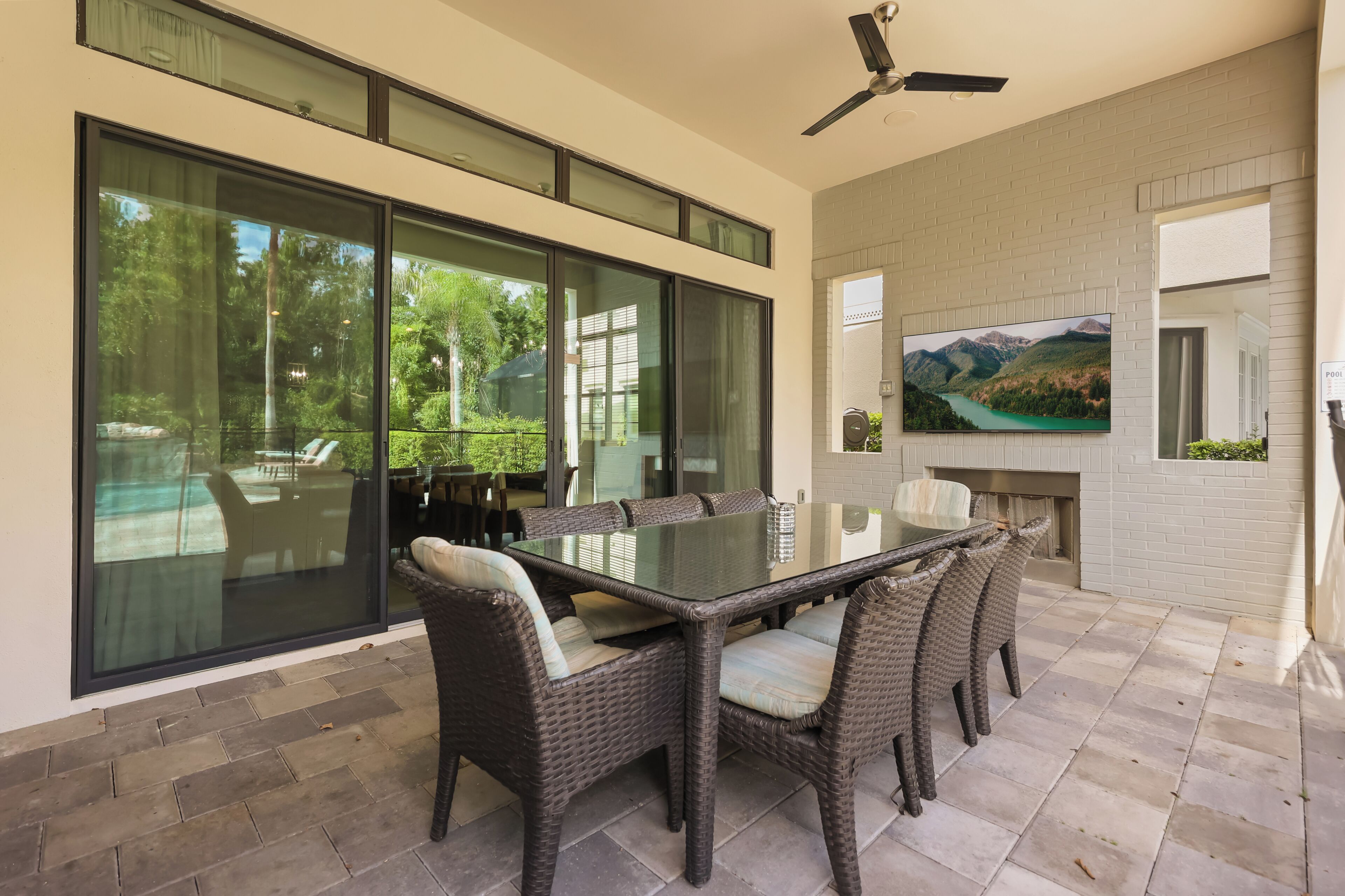 A modern outdoor dining area with a glass-top table and wicker chairs, a ceiling fan, and a TV mounted on the wall.
