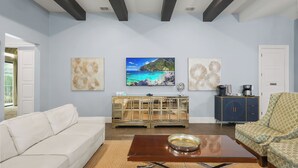 Uma sala de estar com um sofá branco, uma mesa de centro de vidro e uma pintura de uma cena de praia.