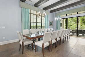 Uma sala de jantar com uma mesa comprida posta para seis pessoas, rodeada por cadeiras, com um candelabro acima e uma grande janela com cortinas.