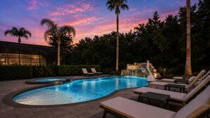 Piscina com espreguiçadeiras e uma cascata, cercada por palmeiras e um edifício com janelas iluminadas.