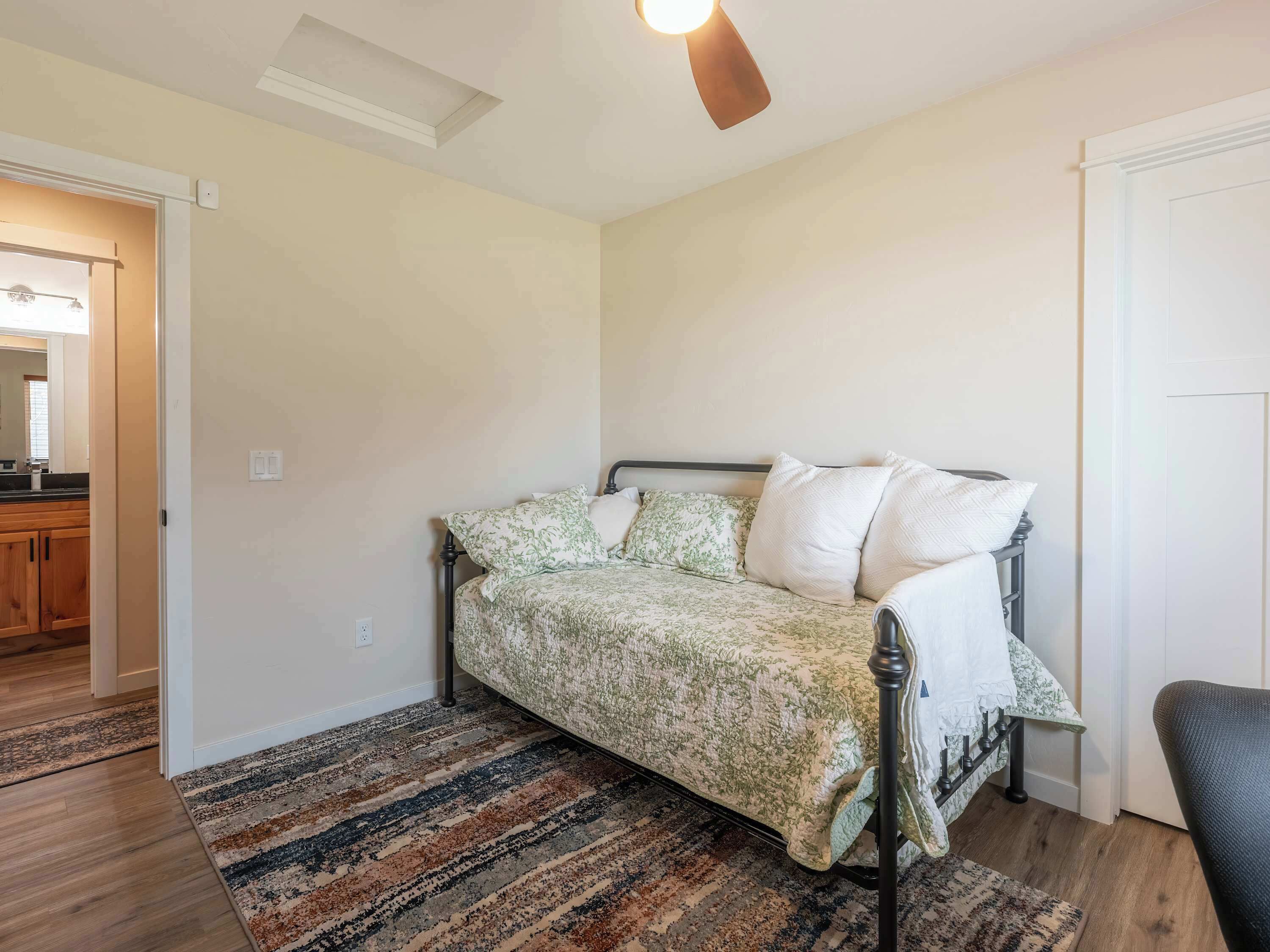 A bedroom with a bed, a patterned rug, a ceiling fan, and a door leading to a bathroom.
