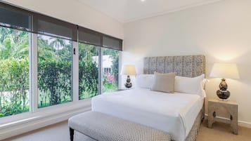 A bedroom with a large bed, two bedside tables with lamps, a bench, and a view of greenery through large windows.