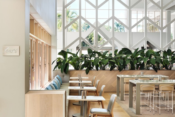 Ein modernes Restaurantambiente mit hohen Decken, großen Fenstern und verschiedenen Sitzgelegenheiten, darunter Tische und Bänke.