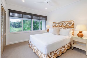 A bedroom with a large bed, two bedside tables, a window with a view of greenery, and a ceiling fan.