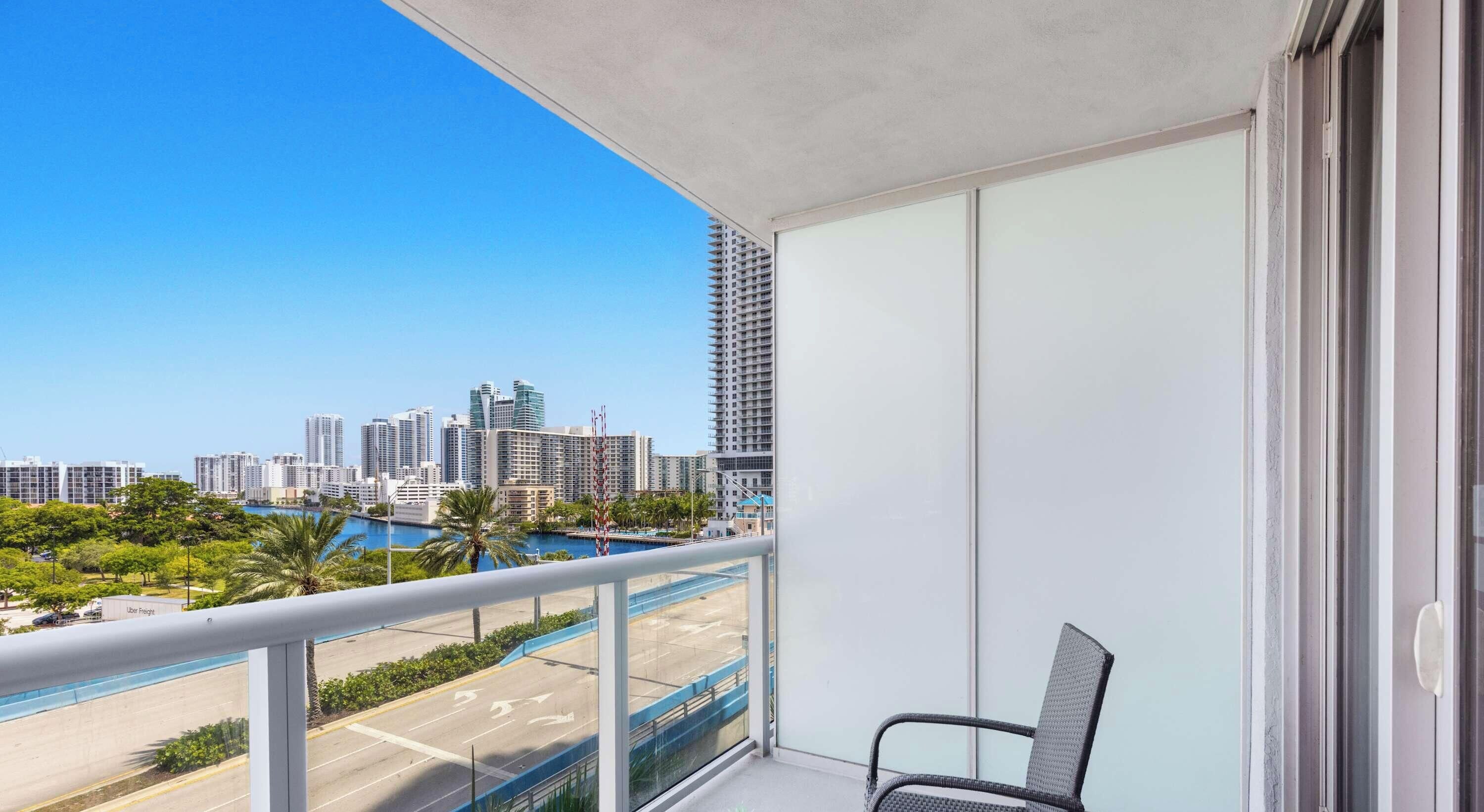 A balcony with a view of a city skyline, a pool, and a chair.