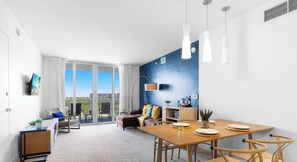 A modern living room with a dining area, large windows, and a blue accent wall. - Bay View 1BR at Beachwalk Elite Resort (Hallandale Beach)