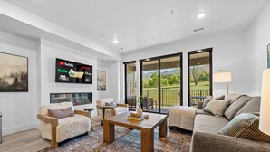 A modern living room with a flat-screen TV displaying streaming services, a fireplace, a sofa, a coffee table, and a view of the outdoors.
