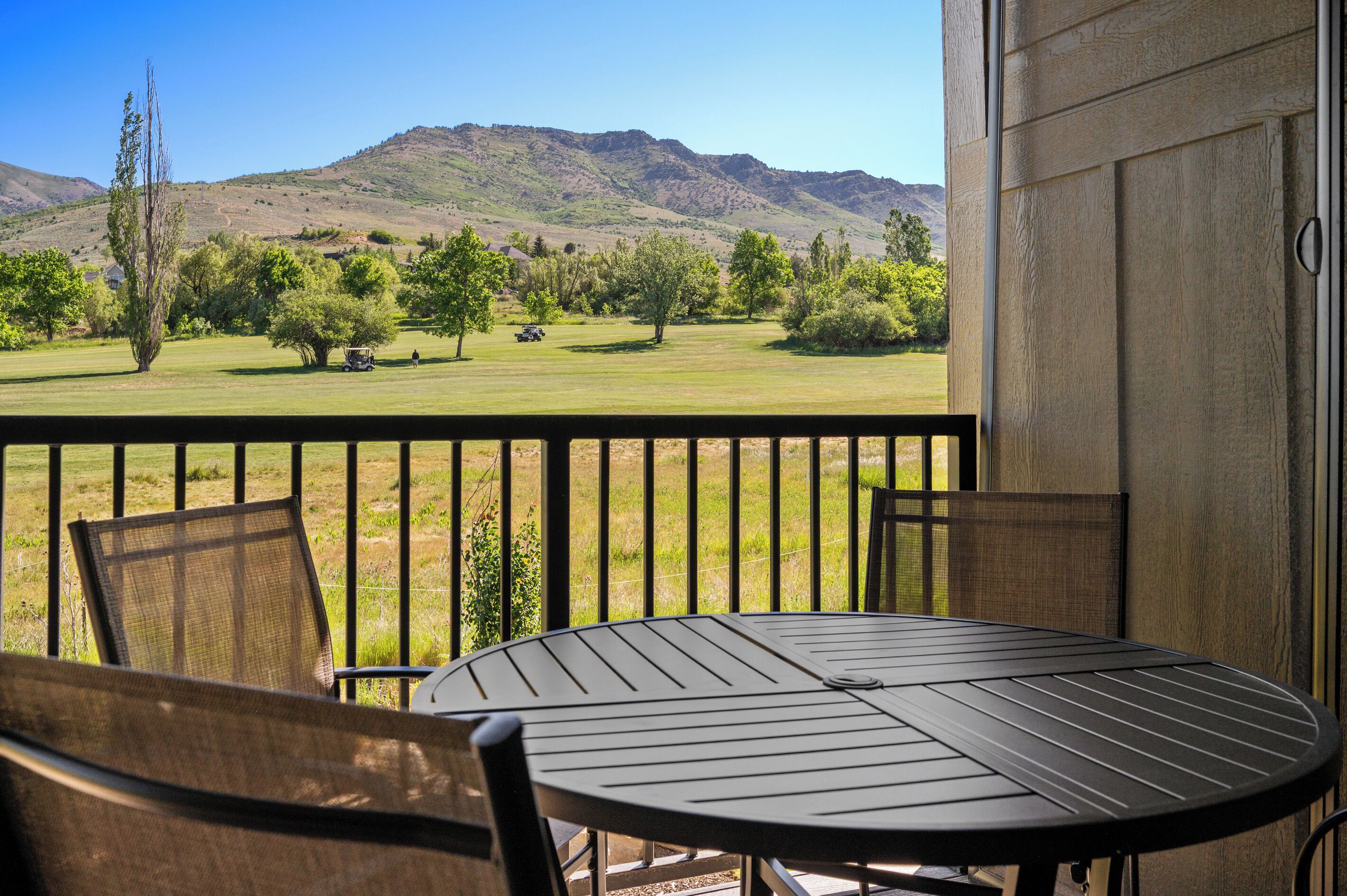 A balcony with a table and chairs, overlooking a grassy field with trees and a mountain in the background.
