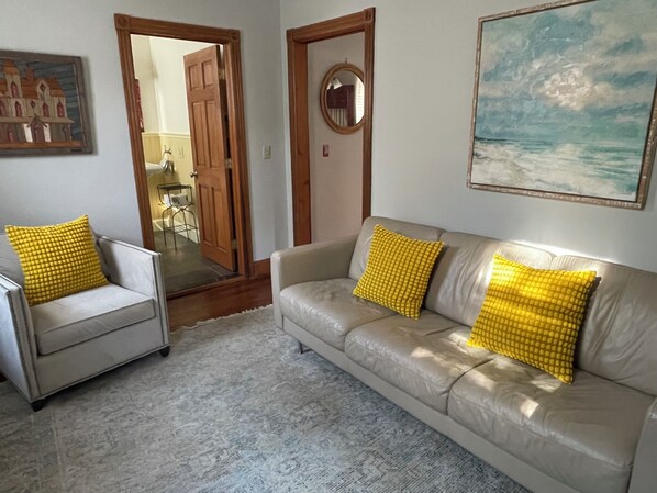 A living room with a sofa and armchair, both with yellow cushions, a wooden door, a round mirror, and a painting on the wall. - Beautiful Ocean Grove Summer & Off-Season Rental (Neptune Township)