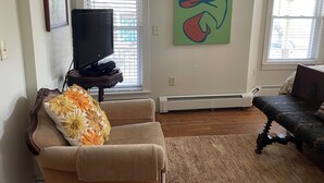Uma sala de estar com uma televisão em um suporte, um sofá com almofada florida, um quadro na parede e uma janela com persianas.