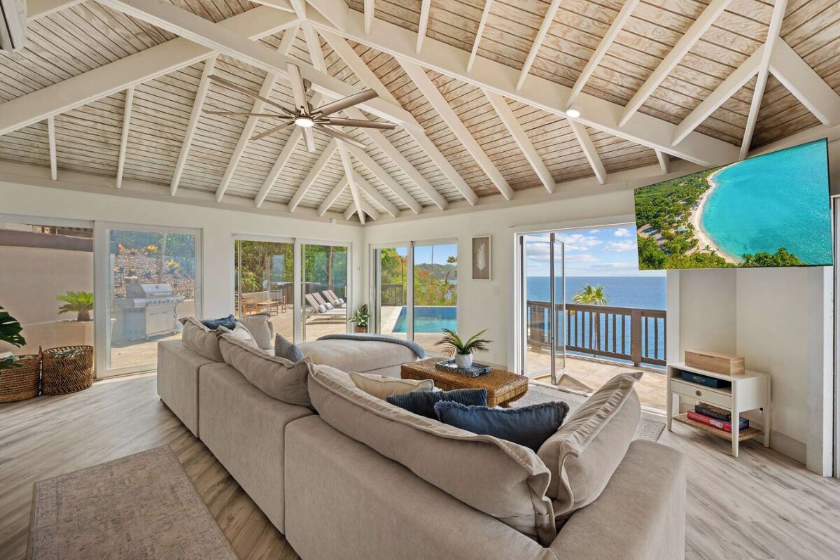 A spacious living room with a large sofa, a ceiling fan, and a TV displaying a beach view.