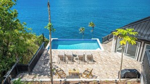 A pool with lounge chairs and a table, overlooking a large body of water and a distant island. - The Parrot's Perch- New 6 BDRM Oceanfront Estate with Pool/Playground/Firepit (Lovenlund)