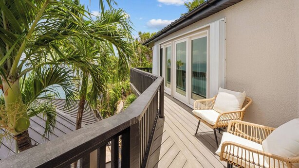 A balcony with wicker chairs, a wooden deck, and a view of palm trees. - The Parrot's Perch- New 6 BDRM Oceanfront Estate with Pool/Playground/Firepit (Lovenlund)
