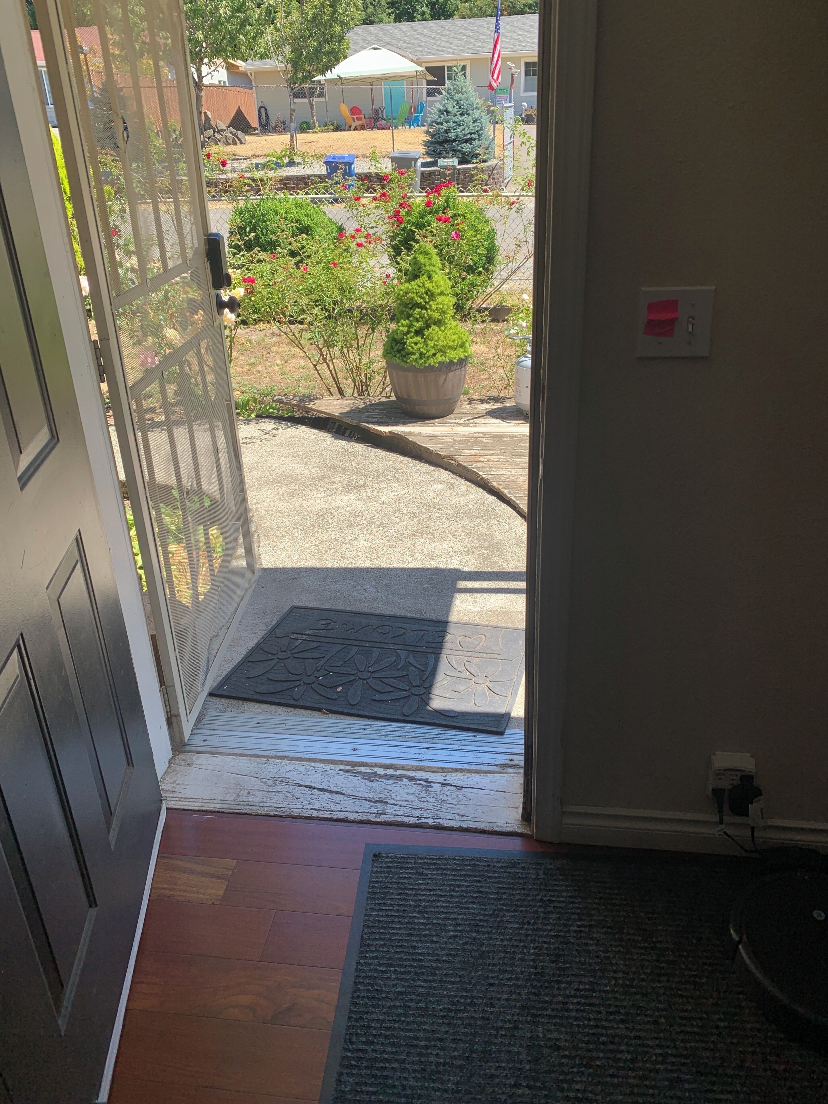 A doorway leading to a covered outdoor area with a doormat, a small garden with potted plants, and a visible American flag.