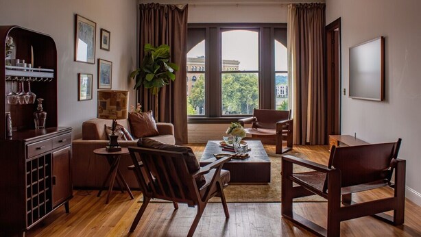 A living room with a sofa, chairs, a coffee table, and a television. There is a window with curtains, a plant, and a view of buildings outside. - The Ilium Suite 401 - Luxury 2 bd Suite in Downtown Troy (Troy)
