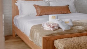 A wooden bed with white bedding, a brown pillow, and a woven bench in a room with a white brick wall and a framed picture above the bed. - The Ilium Suite 401 - Luxury 2 bd Suite in Downtown Troy (Troy)