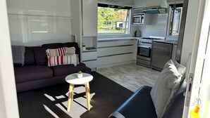 A compact living space with a sofa, a small table, a kitchenette, and a window with a view of greenery.