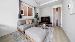 A modern living room with a sofa, ottoman, coffee table, and a TV displaying 'NETFLIX'. - GRACE HOME-A (Sapporo)
