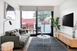 Ein modernes Wohnzimmer mit einem grauen Sofa, einem Couchtisch, einem Fernseher und einem großen Fenster mit Blick auf ein Gebäude mit roten Dächern.