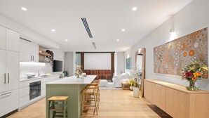 Une cuisine moderne dotée d’un îlot central, de tabourets de bar en bois, d’un four encastré et d’un coin repas avec un canapé blanc et un grand tableau abstrait.